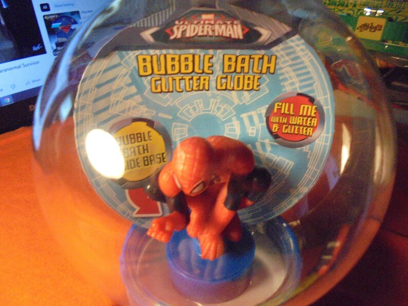 May include: A clear plastic bubble bath glitter globe featuring a red and black Spider-Man figure. The packaging includes text that reads "Bubble Bath Glitter Globe" and "Fill me with water & glitter."