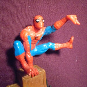 K&ouml;nnte beinhalten: Eine Spider-Man-Actionfigur aus Kunststoff in hockender Pose, die einen rot-blauen Anzug mit einem schwarzen Spinnenemblem auf der Brust tr&auml;gt. Die Figur steht auf einem braunen, kistenartigen Sockel, daneben ist ein gelbes Ma&szlig;band zu sehen.
