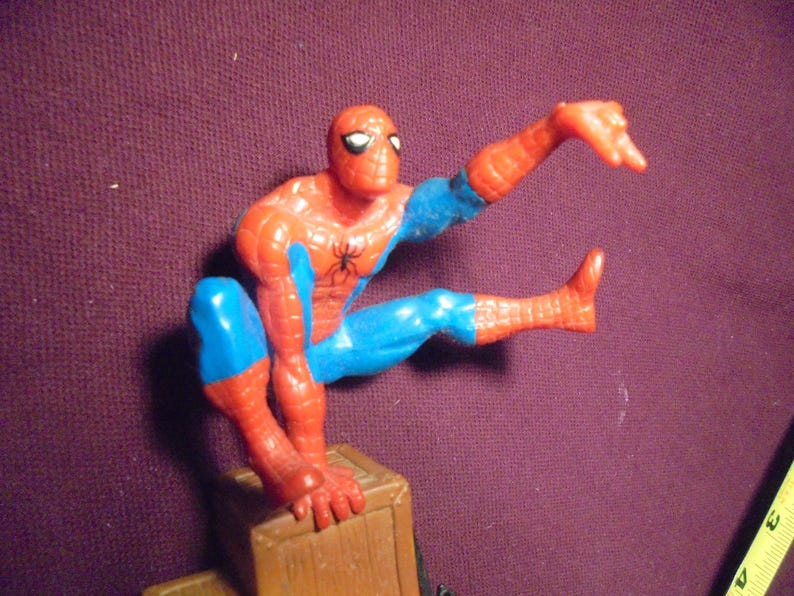 K&ouml;nnte beinhalten: Eine Spider-Man-Actionfigur aus Kunststoff in hockender Pose, die einen rot-blauen Anzug mit einem schwarzen Spinnenemblem auf der Brust tr&auml;gt. Die Figur steht auf einem braunen, kistenartigen Sockel, daneben ist ein gelbes Ma&szlig;band zu sehen.