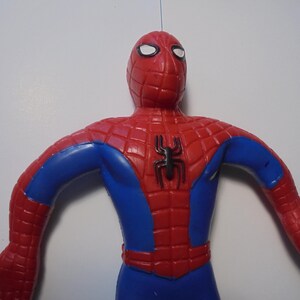 May include: A red and blue Spider-Man action figure featuring a black spider emblem on the chest. The figure has a textured web pattern across the red sections of the head, shoulders, and torso, with white eye lenses and a smooth blue midsection.