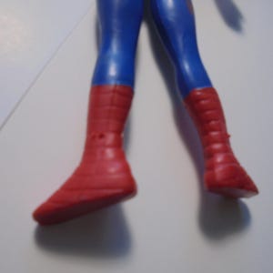 May include: A plastic action figure showing the lower body of a character with blue legs and red boots. The boots feature a textured grid pattern across the surface. The figure is positioned against a plain white background with a partial view of a ruler.
