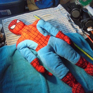 May include: A plush Spider-Man doll in a lying position. The doll features a red and blue color scheme, with a detailed web pattern on the red areas. The eyes are white, and the chest has a black spider emblem. The doll is lying on a blue towel.