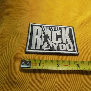 May include: A rectangular black and white patch with the text "WE WILL ROCK YOU" and a silhouette of a person playing a guitar. The patch is approximately 3 inches wide. The patch is on a yellow textured surface.