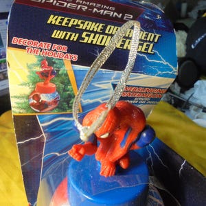 May include: The Amazing Spider-Man 2 keepsake ornament with shower gel inside a blue plastic container. The packaging features lightning graphics, the text "KEEPSAKE ORNAMENT WITH SHOWER GEL," "DECORATE FOR THE HOLIDAYS," and "WEBSLINGING WATERMELON."
