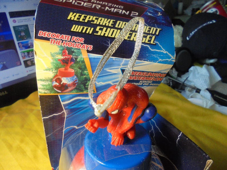 May include: The Amazing Spider-Man 2 keepsake ornament with shower gel inside a blue plastic container. The packaging features lightning graphics, the text "KEEPSAKE ORNAMENT WITH SHOWER GEL," "DECORATE FOR THE HOLIDAYS," and "WEBSLINGING WATERMELON."
