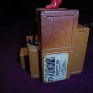 K&ouml;nnte beinhalten: Ein brauner Spielzeugsockel aus Kunststoff in Form eines Stapels Holzkisten mit einem schwarzen Haken an der Seite. Ein wei&szlig;er rechteckiger Aufkleber auf der Vorderseite zeigt einen Strichcode und den Text APPLAUSE INC. WOODLAND HILLS CA 91365 TM & &copy; 1987 MARVEL.