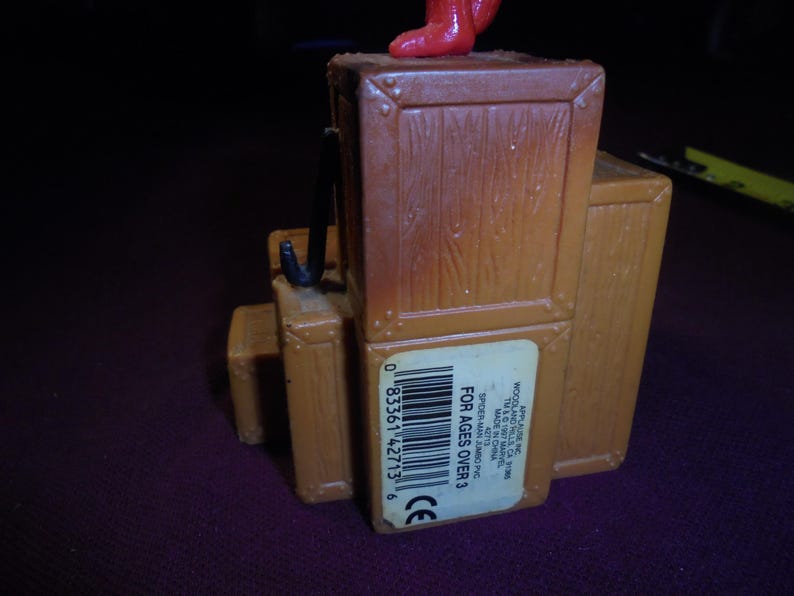 K&ouml;nnte beinhalten: Ein brauner Spielzeugsockel aus Kunststoff in Form eines Stapels Holzkisten mit einem schwarzen Haken an der Seite. Ein wei&szlig;er rechteckiger Aufkleber auf der Vorderseite zeigt einen Strichcode und den Text APPLAUSE INC. WOODLAND HILLS CA 91365 TM & &copy; 1987 MARVEL.