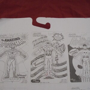 May include: A white cardboard backing featuring three Marvel Super-Hero action figure designs. The figures are The Amazing Spider-Man, Captain America, and The Punisher. Each figure is outlined in black and labeled with the character's name.