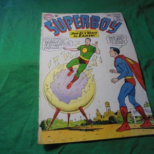 May include: Vintage Superboy comic book cover featuring two costumed characters. One is in a green and red outfit, emerging from a yellow portal. The other is in a blue and red suit with a red cape. The title "Superboy" is at the top.
