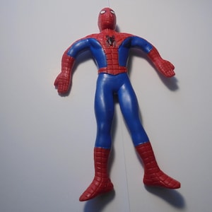 May include: A red and blue Spider-Man action figure with a black spider emblem on the chest. The figure features a web pattern on the red sections of the head, shoulders, and boots. A yellow measuring tape is visible in the upper right corner.