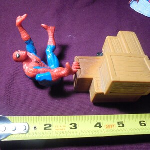 K&ouml;nnte beinhalten: Eine Spider-Man-Actionfigur in einem rot-blauen Anzug neben einem Stapel beiger Kunststoffkisten. Ein gelbes Ma&szlig;band liegt im Vordergrund und zeigt, dass die Kisten eine Gesamtl&auml;nge von etwa 11,4 cm aufweisen.