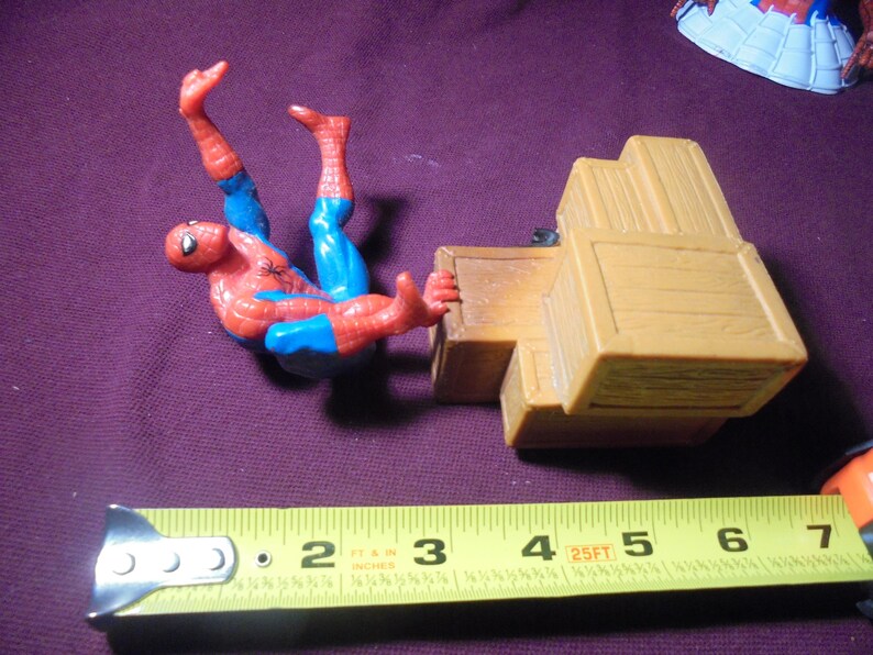 K&ouml;nnte beinhalten: Eine Spider-Man-Actionfigur in einem rot-blauen Anzug neben einem Stapel beiger Kunststoffkisten. Ein gelbes Ma&szlig;band liegt im Vordergrund und zeigt, dass die Kisten eine Gesamtl&auml;nge von etwa 11,4 cm aufweisen.