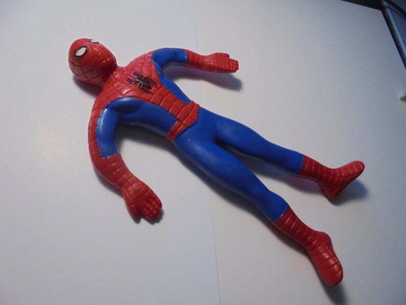 May include: A red and blue Spider-Man action figure with a web pattern on the head, chest, arms, and legs. The figure features a black spider emblem on the chest and white eye lenses. The toy is posed with arms and legs extended in a flat position.