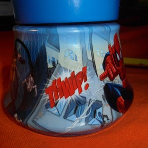 May include: A blue and white tumbler featuring Spider-Man in action. The design includes comic book style graphics of buildings and the word "THWIP!" in red. The tumbler has a blue lid.