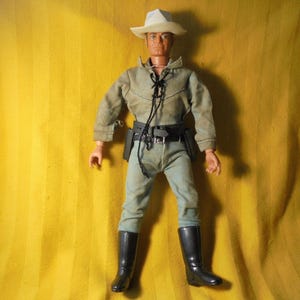 May include: A vintage cowboy action figure, wearing a light green shirt, pants, and a white hat. The figure has a black belt with holsters and black boots. The figure is standing against a yellow background.