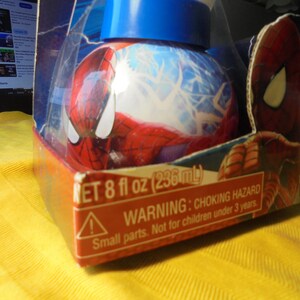 May include: A round Spider-Man themed container with a blue lid, encased in a cardboard box featuring Spider-Man graphics. The packaging displays the text NET 8 fl oz (236 mL) and a warning about a choking hazard for children under 3 years.