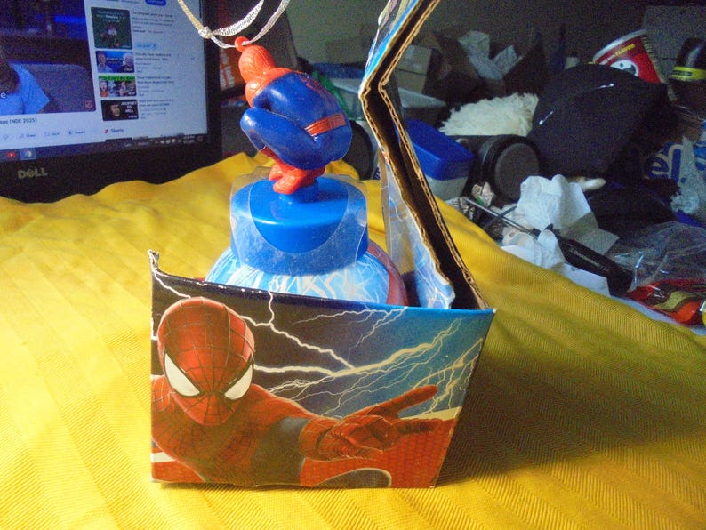 May include: A Spider-Man themed plastic water bottle housed in a cardboard display box. The bottle features a blue cap topped with a small red and blue Spider-Man figurine. The box displays a graphic of Spider-Man with white lightning bolts.