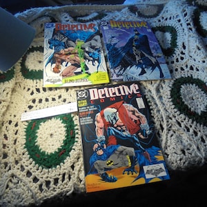 May include: Three vintage comic books with the title "Detective Comics" on the cover. The comic books are on a white crocheted blanket. The comic books are in good condition and have colorful covers.