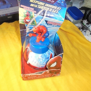 May include: A Spider-Man 2 keepsake ornament with shower gel in a blue and red box. The packaging features the text Keepsake Ornament with Shower Gel, Webslinging Watermelon, and 8 fl oz (236 ml). A small Spider-Man figure sits atop the blue lid.