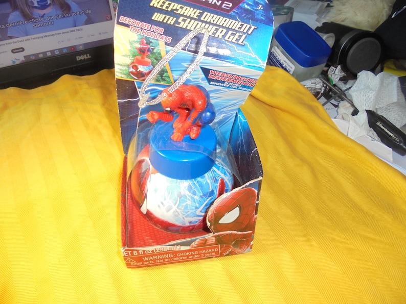 May include: A Spider-Man 2 keepsake ornament with shower gel in a blue and red box. The packaging features the text Keepsake Ornament with Shower Gel, Webslinging Watermelon, and 8 fl oz (236 ml). A small Spider-Man figure sits atop the blue lid.