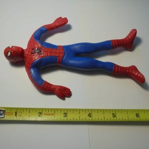 May include: A red and blue Spider-Man action figure with a black spider emblem on the chest. The figure is positioned above a yellow measuring tape, which indicates that the toy measures approximately 7 inches in total length from head to toe.