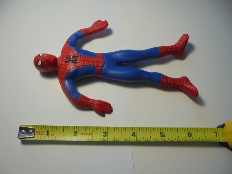 May include: A red and blue Spider-Man action figure with a black spider emblem on the chest. The figure is positioned above a yellow measuring tape, which indicates that the toy measures approximately 7 inches in total length from head to toe.