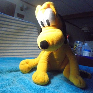 May include: A plush toy of Pluto, a yellow dog with black ears and a black nose, sits on a blue towel. The toy has large white eyes and is in a sitting position. The background includes a striped blanket and various items.