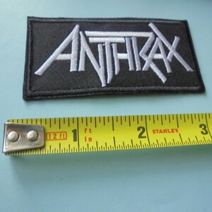 May include: Black rectangular patch with the word "ANTHRAX" embroidered in white, stylized lettering. A yellow tape measure is placed below the patch, showing measurements in inches.