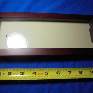 May include: A rectangular, dark-stained wooden display case with a glass front. The interior has a cream-colored backing. A yellow measuring tape is placed below the case, showing measurements in inches.