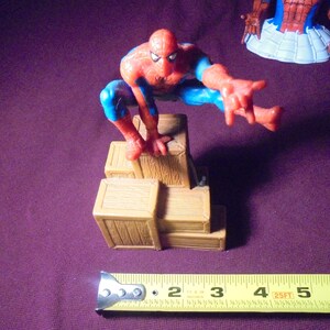 K&ouml;nnte beinhalten: Eine rot-blaue Spider-Man-Figur in hockender Pose, die auf einem Stapel brauner Holzkisten thront. Ein gelbes Ma&szlig;band liegt im Vordergrund und zeigt, dass die Figur eine Gesamth&ouml;he von etwa 14 cm aufweist.