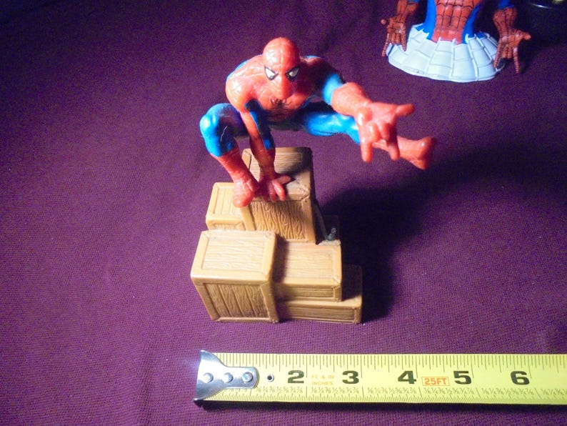 K&ouml;nnte beinhalten: Eine rot-blaue Spider-Man-Figur in hockender Pose, die auf einem Stapel brauner Holzkisten thront. Ein gelbes Ma&szlig;band liegt im Vordergrund und zeigt, dass die Figur eine Gesamth&ouml;he von etwa 14 cm aufweist.