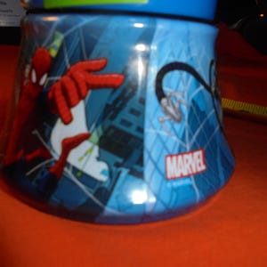 May include: A blue Marvel-branded tumbler featuring Spider-Man. The design includes Spider-Man in action with red and white accents against a cityscape backdrop. The tumbler has a blue lid and a curved shape.