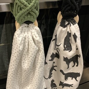 Könnte beinhalten: Zwei Geschirrtuchhalter. Einer ist olivgrün, der andere schwarz. Jeder hat einen Holzring und ein gemustertes Handtuch. Eines hat einen weißen Hintergrund mit grauen Strichen. Das andere hat schwarze Katzen-Silhouetten.