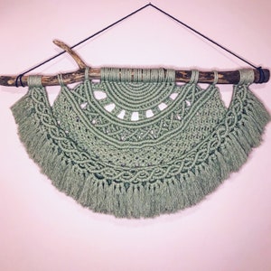 May include: Sage green macrame wall hanging. The handmade piece features a half-circle design with intricate knotting and fringe. It is suspended from a natural wooden branch and black cord, adding a touch of bohemian decor to any space.