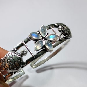 Può includere: Bracciale a polsino argentato con un design floreale con pietre iridescenti e due dettagli di tartarughe. Il bracciale aperto misura circa 6,35 cm di diametro, con un elemento centrale floreale e dettagli di tartarughe.