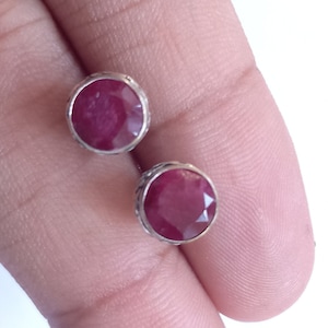 Puede incluir: Un par de pendientes de botón redondos de color rubí. Cada pendiente presenta una piedra preciosa facetada de color rojo intenso engastada en un bisel plateado. Los pendientes se muestran sobre un fondo claro.