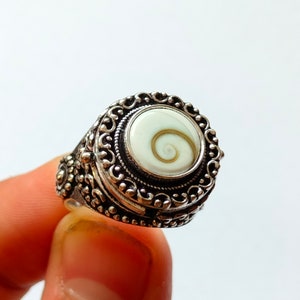 May include: A silver ring with an ornate design and a white shell inlay. The shell has a spiral pattern.