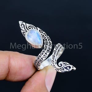 May include: A silver-toned ring with a teardrop-shaped, iridescent gemstone. The ring has an ornate, swirling design with the text "Megharrration5" visible. The gemstone appears to be a moonstone, with a pearlescent, blue and white color.