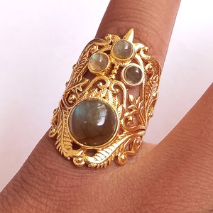 Può includere: Un anello d'oro con una grande pietra di labradorite grigio scuro circondata da quattro pietre più piccole e trasparenti. L'anello ha un design dettagliato e ornato con motivi a forma di foglie.
