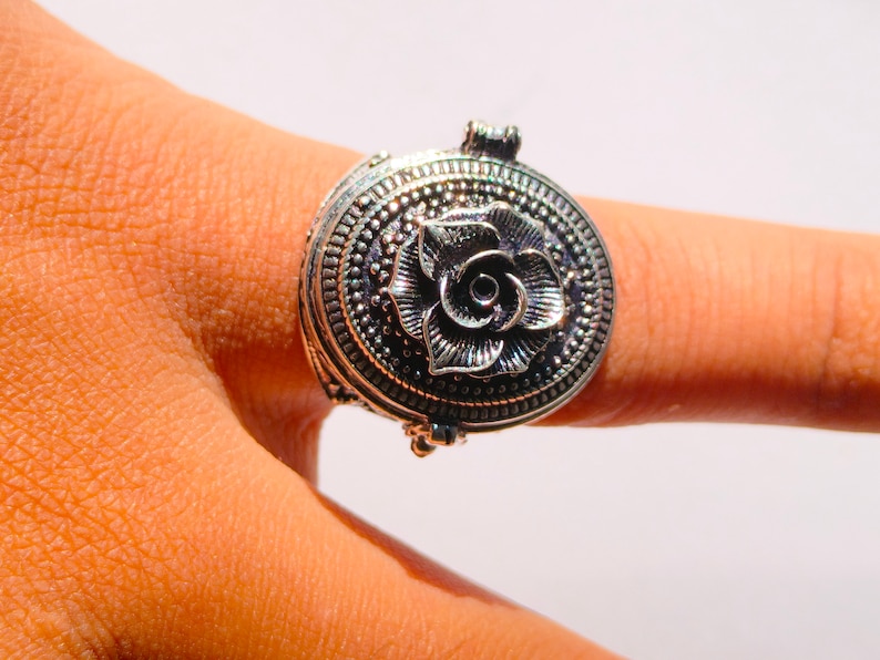 K&ouml;nnte beinhalten: Ein silberner Ring mit einem ovalen, klappbaren Anh&auml;nger mit einem detaillierten Rosenmotiv.