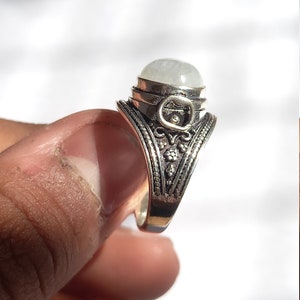 May include: A silver ring with an oval-shaped white gemstone set in a decorative silver bezel. The ring band is intricately detailed with floral and geometric patterns.