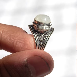 May include: A silver ring with an oval-shaped white stone set in the center. The ring has intricate floral designs and a hinged lid on the stone.