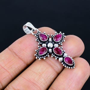 May include: A silver cross pendant with five deep red gemstones. The teardrop-shaped stones are set within a detailed silver frame, with small silver beads. A loop is at the top for a chain.