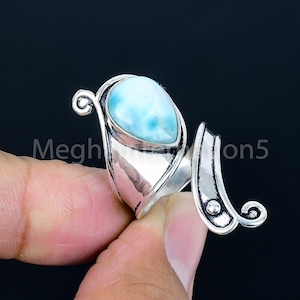 Puede incluir: Un anillo de plata con una piedra preciosa azul claro en forma de pera. El anillo tiene un diseño abierto con remolinos decorativos y el texto "Megh" y "on5". La piedra preciosa está engastada en un bisel.