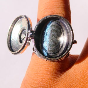 K&ouml;nnte beinhalten: Ein silberner Ring mit einem runden, klappbaren Anh&auml;nger. Der Anh&auml;nger ist ge&ouml;ffnet und das Innere ist sichtbar. Der Ring befindet sich an einem Finger.