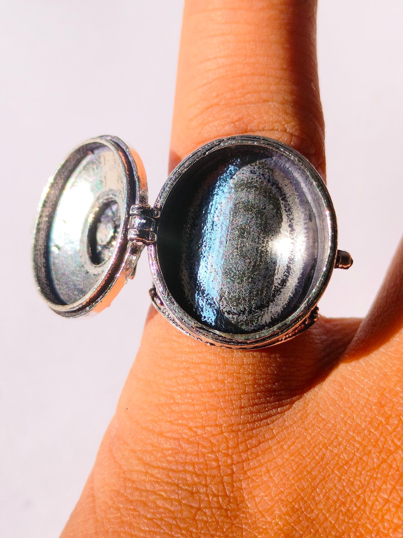K&ouml;nnte beinhalten: Ein silberner Ring mit einem runden, klappbaren Anh&auml;nger. Der Anh&auml;nger ist ge&ouml;ffnet und das Innere ist sichtbar. Der Ring befindet sich an einem Finger.