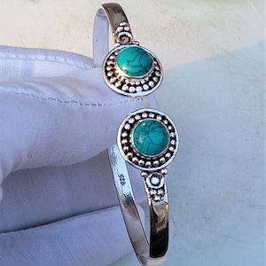 May include: A silver-colored cuff bracelet with two circular turquoise-colored stones. Each stone is surrounded by a beaded border. The bracelet is open, with the ends curving slightly away from each other.