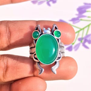 Könnte beinhalten: Ein silberner Ring mit einem grünen Edelstein in der Mitte und zwei kleineren grünen Edelsteinen an beiden Seiten. Der Ring ist so gestaltet, dass er wie ein Insekt mit Beinen aussieht.