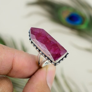 May include: A silver ring featuring a large, rectangular, faceted ruby gemstone. The ruby is a deep, vibrant red color. The ring has a detailed, beaded border around the gemstone and a split shank design.