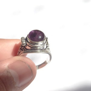 May include: A silver ring featuring a large, round, deep purple gemstone. The ring has an ornate, detailed band with decorative elements. The gemstone is set in a bezel setting, and the ring is held by a hand.